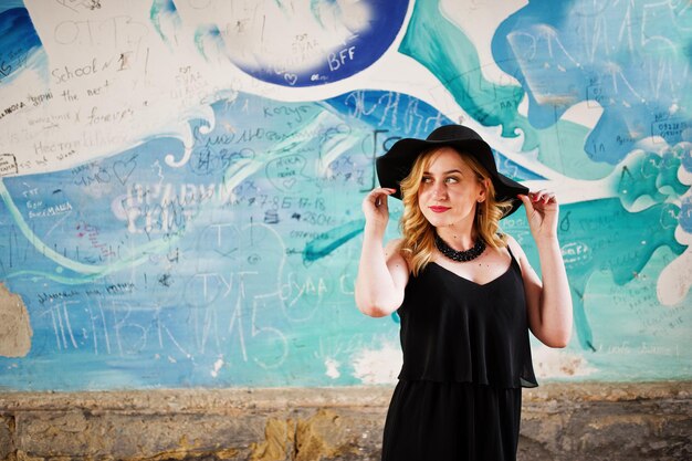 Blonde Frau auf schwarzen Kleiderhalsketten und Hut gegen Graffitiwand