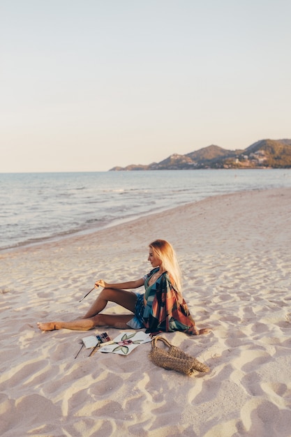 Blonde Frau, die Aquarellblume durch Pinsel auf dem Strand zeichnet