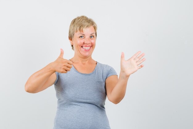 Blonde Frau, die Daumen oben im hellblauen T-Shirt zeigt und fröhlich schaut. Vorderansicht.