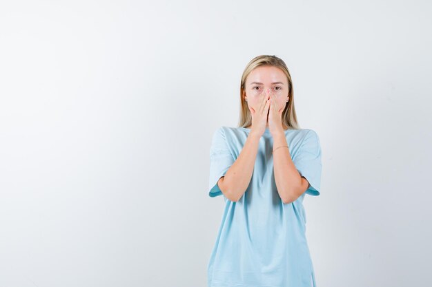 Blonde Frau, die den Mund mit den Händen im blauen T-Shirt bedeckt und überrascht schaut