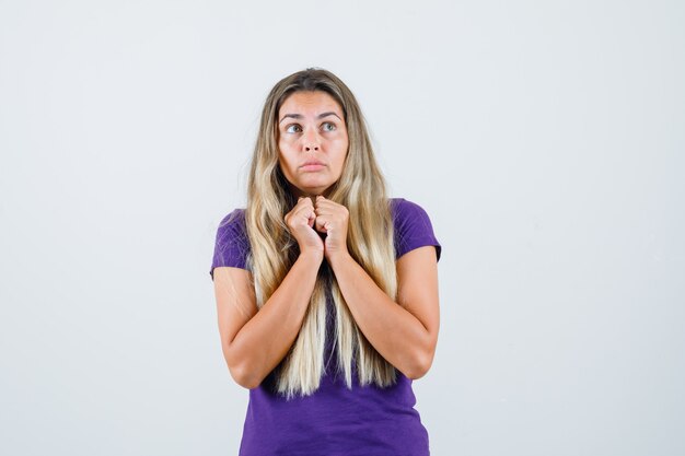 blonde Frau, die Fäuste zusammen auf Brust in violettem T-Shirt hält und ängstlich schaut, Vorderansicht.