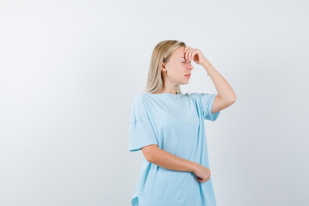 Blonde Frau, die Hand auf dem Kopf hält, Kopfschmerzen in blauem T-Shirt hat und erschöpft aussieht