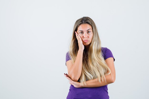 blonde Frau, die Handfläche auf Wange im violetten T-Shirt hält und nachdenklich schaut. Vorderansicht.