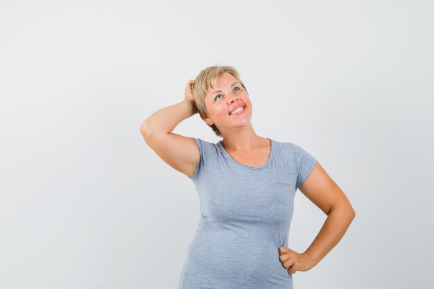 Blonde Frau, die ihre Hand auf Taille legt, über etwas nachdenkt und in hellblauem T-Shirt nach oben schaut und nachdenklich aussieht. Vorderansicht.