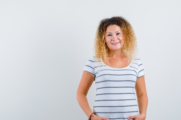 Blonde Frau, die im gestreiften T-Shirt die Hände auf den Hüften hält und fröhlich aussieht. Vorderansicht.