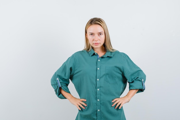 Blonde Frau, die im grünen Hemd die Hände an der Taille hält und vernünftig aussieht looking