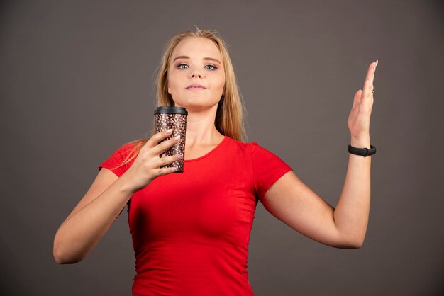 Kostenloses Foto blonde frau, die kaffee zum mitnehmen an schwarzer wand hält.