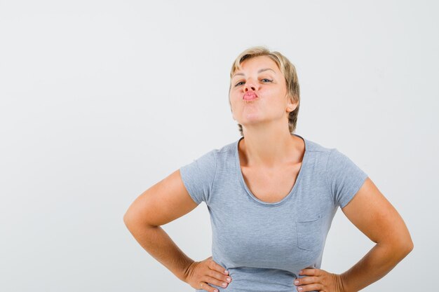 Blonde Frau, die Küsse sendet und ihre Hände auf Taille im hellblauen T-Shirt hält und fröhlich aussieht. Vorderansicht.