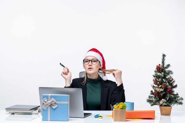 Blonde Frau, die mit einem Weihnachtsmannhut spielt, der an einem Tisch mit einem Weihnachtsbaum und einem Geschenk sitzt