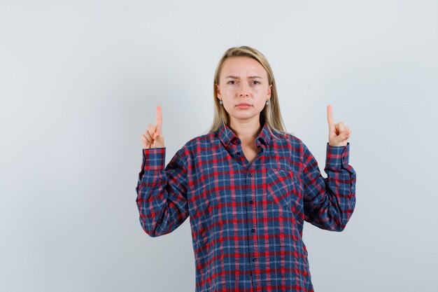 Blonde Frau, die mit Zeigefingern im karierten Hemd nach oben zeigt und wütend aussieht. Vorderansicht.