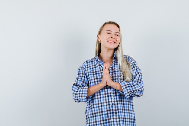 Blonde Frau, die namaste Geste im blauen karierten Hemd des Gingham zeigt und strahlend schaut. Vorderansicht.