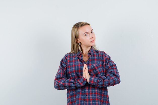 Blonde Frau, die namaste Geste im karierten Hemd zeigt und strahlende Vorderansicht schaut.