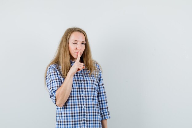 Blonde Frau, die Stille Geste zeigt, Auge im Hemd zwinkert und selbstbewusst aussieht.