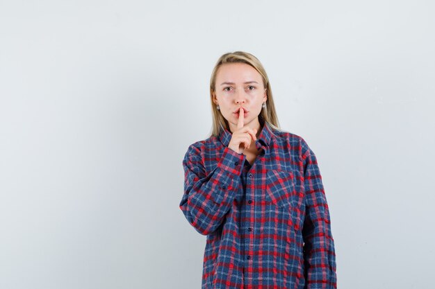 Blonde Frau, die Stille Geste zeigt, Zeigefinger auf Mund in kariertem Hemd legt und konzentriert schaut. Vorderansicht.