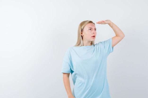 Blonde Frau, die weit weg mit der Hand über dem Kopf in blauem T-Shirt schaut und konzentriert aussieht