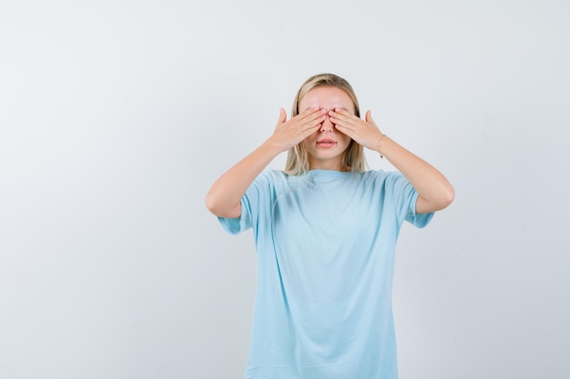 Blonde Frau im blauen T-Shirt, die die Augen mit den Händen bedeckt und ernst schaut