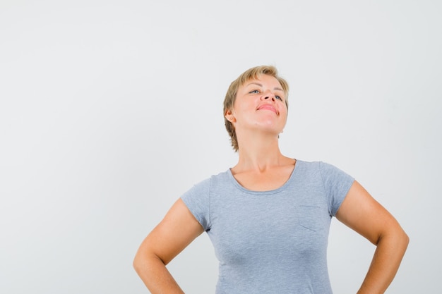 Blonde Frau im hellblauen T-Shirt, das Hände auf Taille hält und Selbstbewusstsein, Vorderansicht aufwirft und schaut.