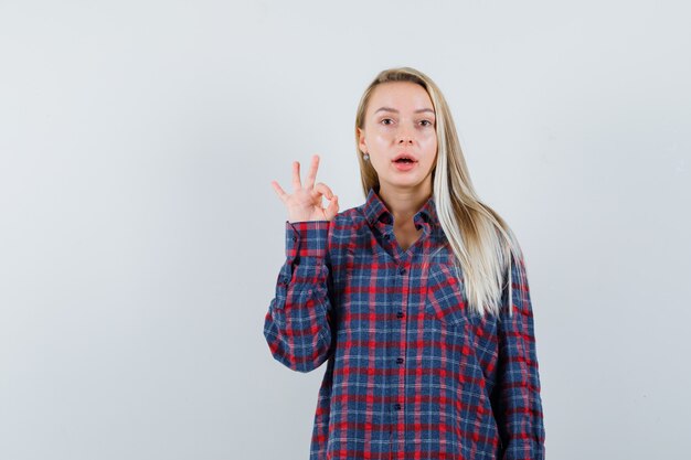 Blonde Frau im karierten Hemd, das ok Zeichen zeigt und attraktive Vorderansicht schaut.