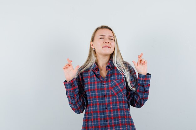 Blonde Frau im karierten Hemd, die Finger gekreuzt hält, Lippen beißt und glücklich schaut, Vorderansicht.