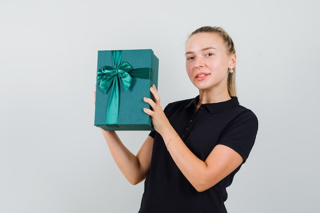 Blonde Frau im schwarzen T-Shirt, das Geschenkbox hält und lächelt und glücklich schaut