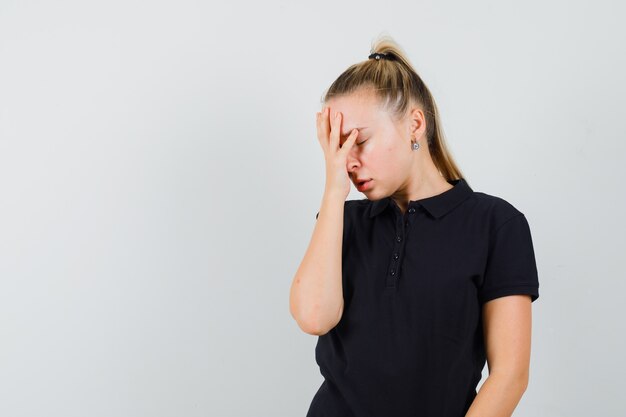 Blonde Frau im schwarzen T-Shirt, das ihre Augen mit den Händen bedeckt und müde aussieht