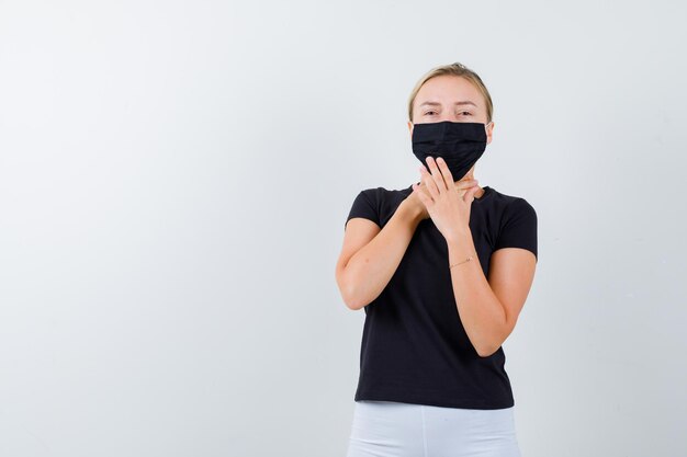 Blonde Frau im schwarzen T-Shirt, weiße Hose, schwarze Maske, die Händchen vor dem Hals hält