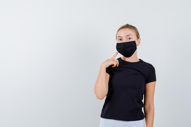 Blonde Frau im schwarzen T-Shirt, weiße Hose, schwarze Maske mit Zeigefinger