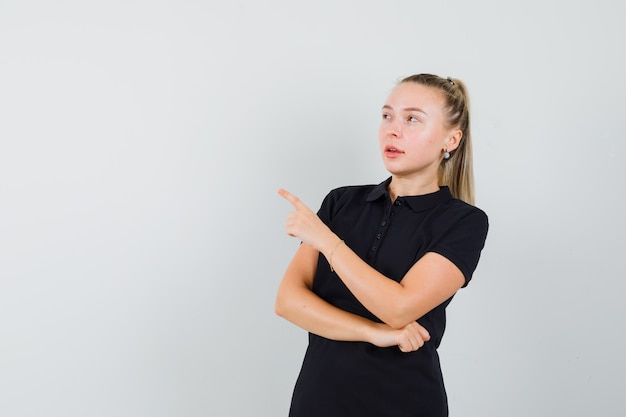 Blonde frau im schwarzen t-shirt zeigt auf die linke seite und lächelt und sieht optimistisch aus