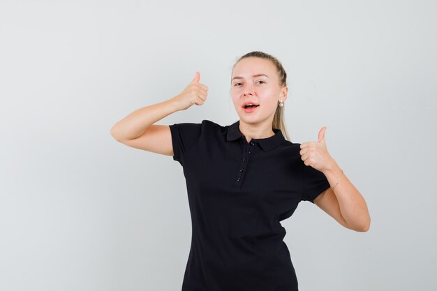 Blonde Frau im schwarzen T-Shirt zeigt Daumen hoch und sieht optimistisch aus