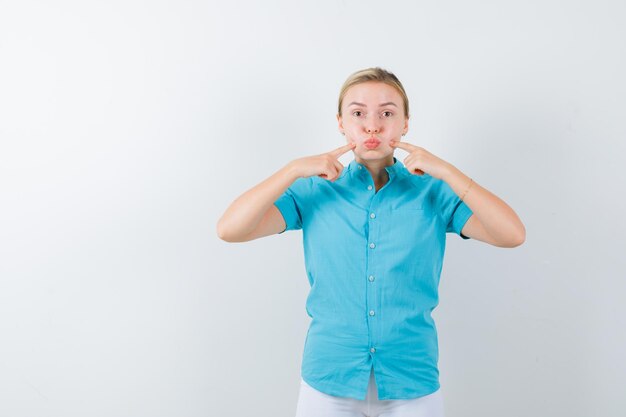 Blonde Frau in blauer Bluse, die Finger auf geschwollenen Wangen hält und isoliert süß aussieht