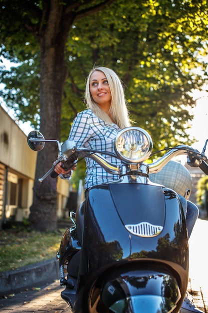 Kostenloses Foto blonde frau in lässiger kleidung, die auf motoroller über grünem baumhintergrund verfügt.
