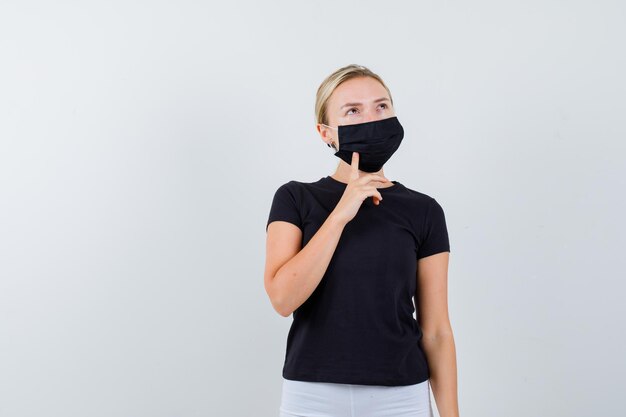 Blonde Frau in schwarzem T-Shirt, weißer Hose, schwarzer Maske mit Zeigefinger am Kinn