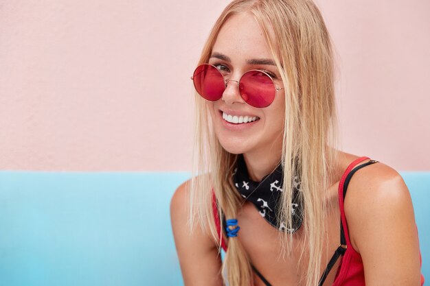 blonde Frau in trendiger Sonnenbrille, trägt modische Kleidung und rote Sonnenbrille, sitzt an der rosa Wand auf bequemer Couch.