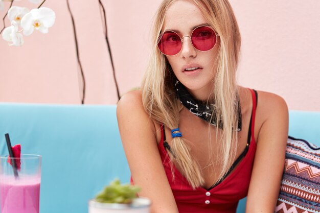 blonde Frau in trendiger Sonnenbrille, trägt modische Kleidung und rote Sonnenbrille, sitzt an der rosa Wand auf bequemer Couch.