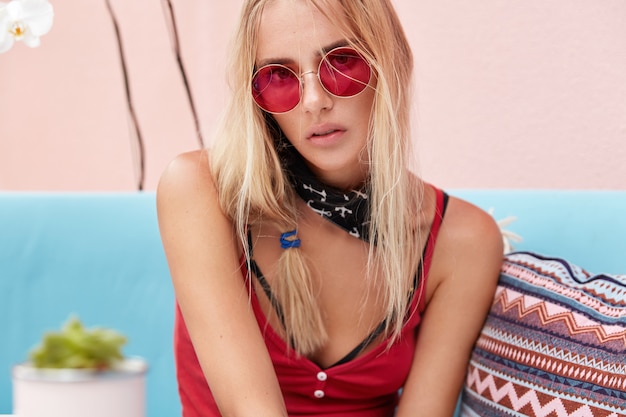 Kostenloses Foto blonde frau in trendiger sonnenbrille, trägt modische kleidung und rote sonnenbrille, sitzt an der rosa wand auf bequemer couch.