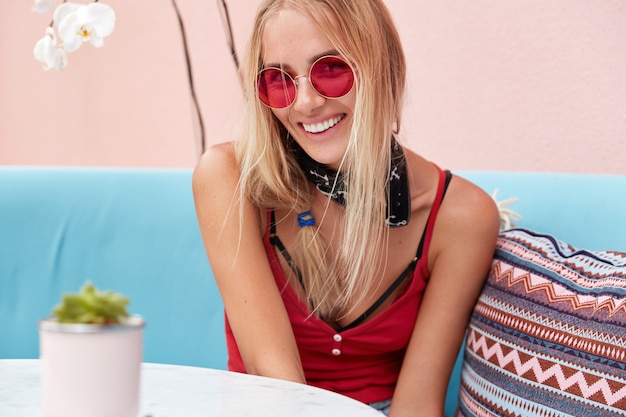 blonde Frau in trendiger Sonnenbrille, trägt modische Kleidung und rote Sonnenbrille, sitzt an der rosa Wand auf bequemer Couch.