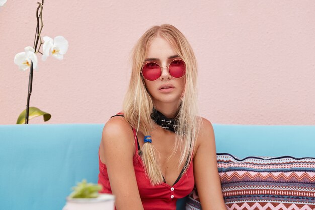 blonde Frau in trendiger Sonnenbrille, trägt modische Kleidung und rote Sonnenbrille, sitzt an der rosa Wand auf bequemer Couch.