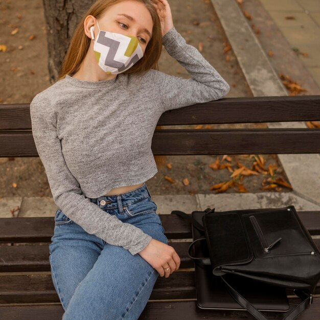 Blonde Frau mit der medizinischen Maske, die auf einer Bank sitzt