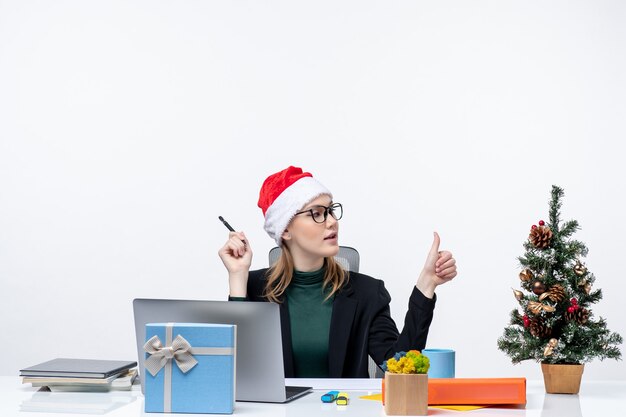 Blonde Frau mit einem Weihnachtsmannhut, der an einem Tisch mit einem Weihnachtsbaum und einem Geschenk sitzt