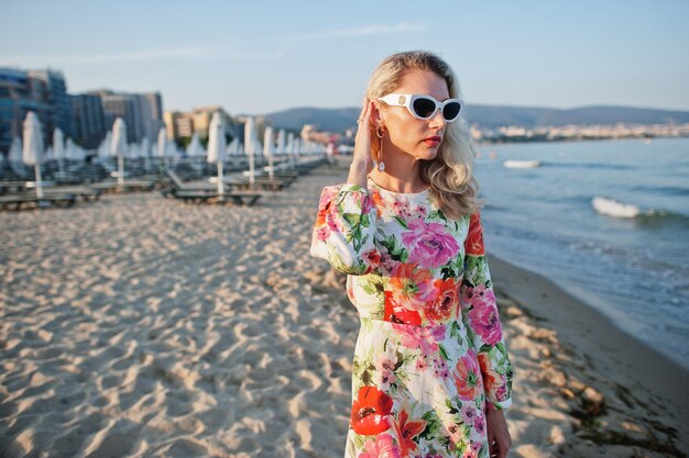 Blonde Frau mit Kleid und Sonnenbrille steht am Sandstrand am Meer und genießt den Blick auf den Sonnenuntergang