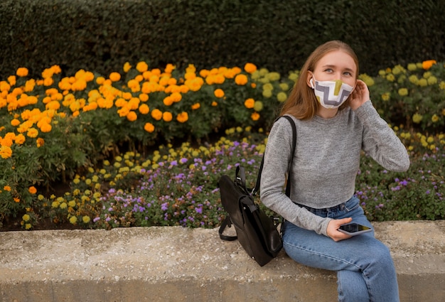 Blonde Frau mit medizinischer Maske, die neben einem Garten mit Kopienraum sitzt