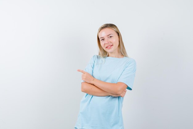 Blonde Frau mit verschränkten Armen, die mit dem Zeigefinger nach links zeigt, im blauen T-Shirt