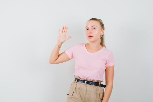 Blonde Frau winkt Hand für Gruß in T-Shirt, Hose und schön aussehende Vorderansicht.