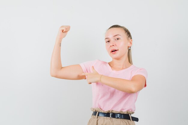 Blonde Frau zeigt auf Armmuskeln in T-Shirt, Hose und sieht selbstbewusst aus. Vorderansicht.