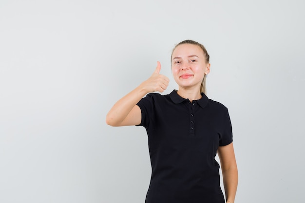 Blonde Frau zeigt Daumen hoch in schwarzem T-Shirt und sieht optimistisch aus