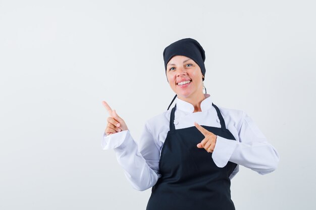 Blonde Frau zeigt nach links mit Zeigefingern in schwarzer Kochuniform und sieht hübsch aus