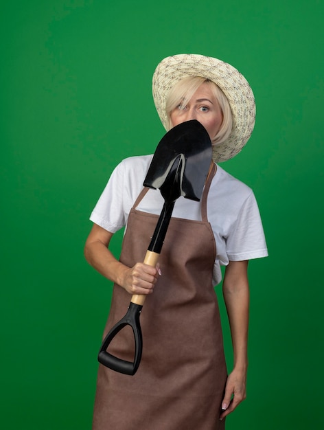 Blonde Gärtnerin mittleren Alters in Uniform mit Hut, der von hinten Spaten hält