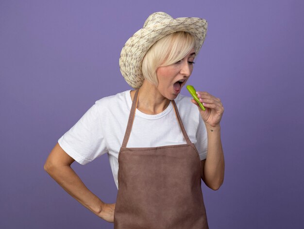Blonde Gärtnerin mittleren Alters in Uniform mit Hut, die Pfeffer in der Nähe des Mundes hält und sie betrachtet, wie sie sich bereit macht, sie zu beißen und die Hand auf der Taille hält