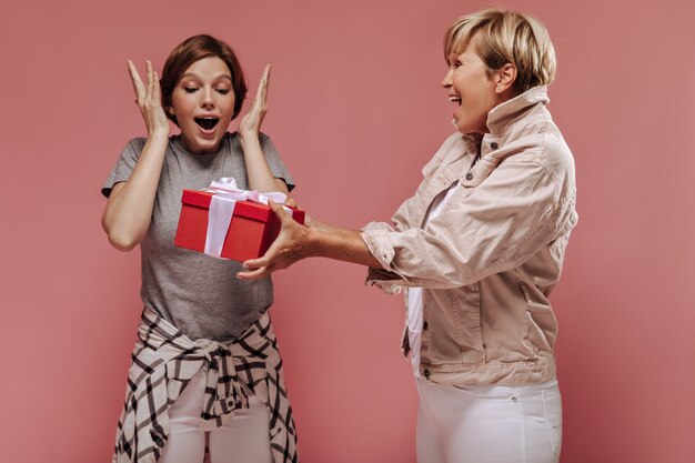 Blonde haarige alte Frau in der Jacke, die aufregendes Mädchen mit kurzer Frisur und kariertem Hemd auf rosa Hintergrund rote Geschenkbox gibt.