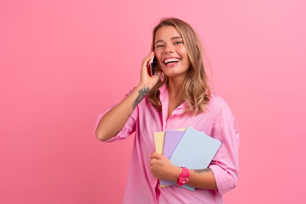 Blonde hübsche Frau im rosa Hemd, die lächelt, hält Notizbücher in der Hand und benutzt das Smartphone, das auf rosa posiert